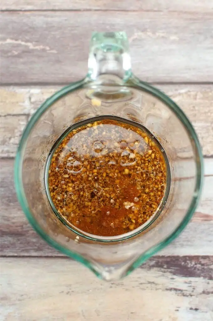 Close-up of a glass measuring cup filled with a savory brown mixture, reminiscent of Mongolian Beef sauce, dotted with seasoning and tiny bubbles on the surface, set against a rustic wooden background.