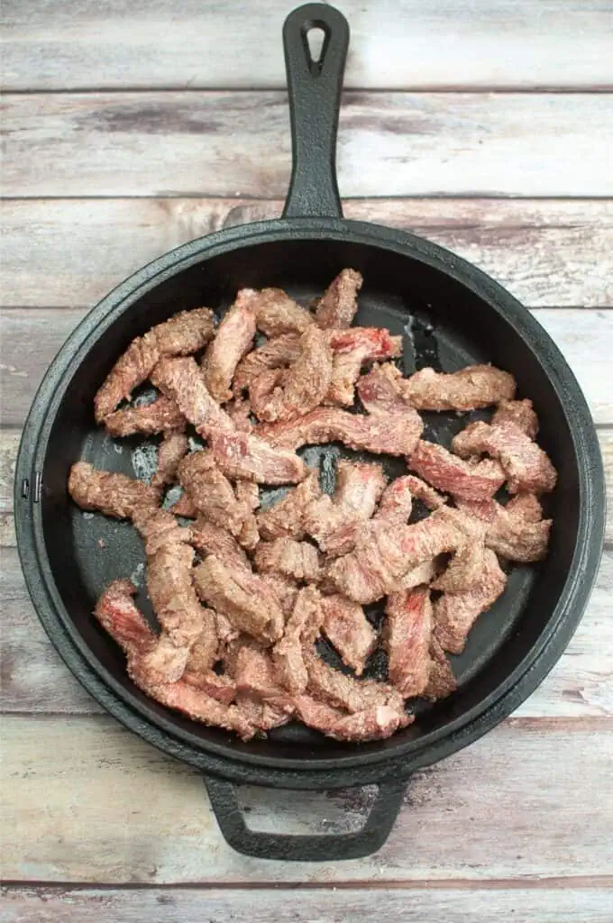 Sliced beef strips sizzle in a black skillet on a rustic wooden surface, capturing the essence of a classic Mongolian Beef Recipe.
