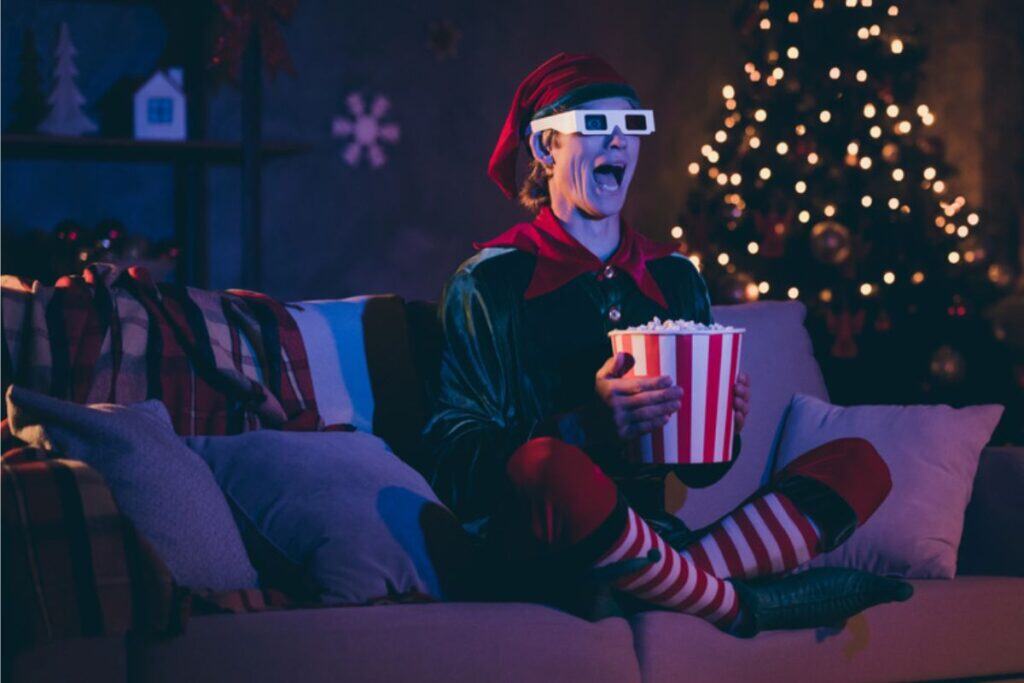 A person dressed as an elf sits on a couch, wearing 3D glasses and holding a large tub of popcorn, enjoying a fun movie night with a decorated Christmas tree in the background.