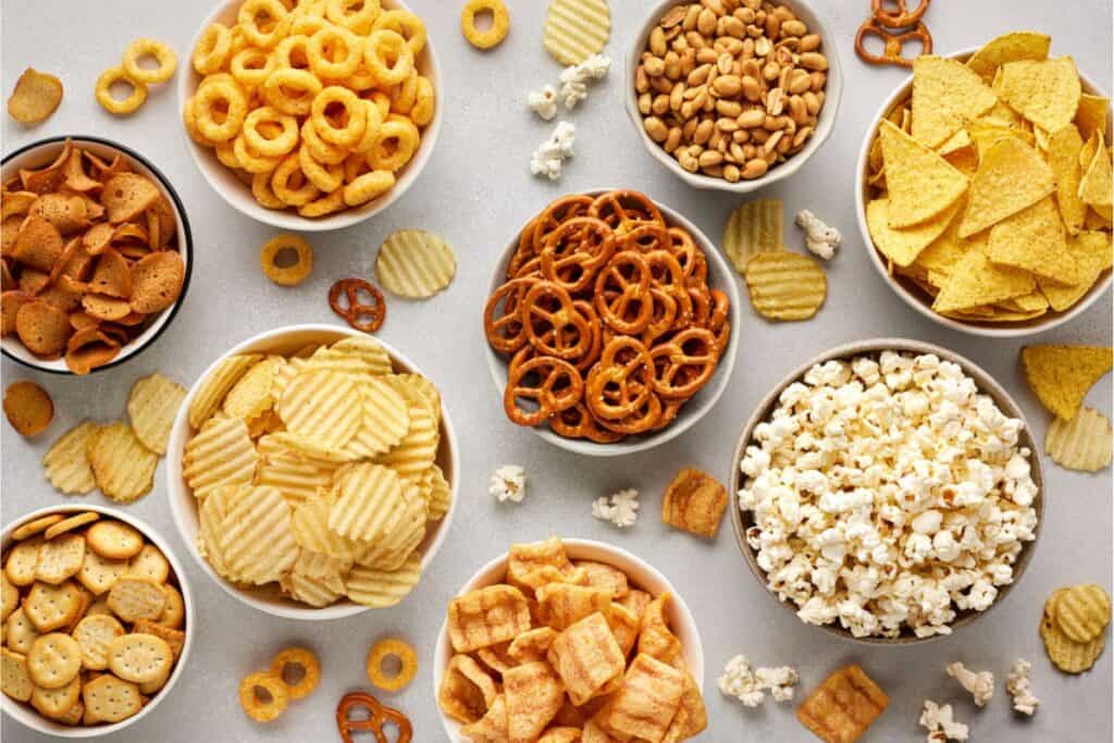 Assorted bowls of snacks, perfect for a fun movie night, including pretzels, popcorn, potato chips, crackers, tortilla chips, nuts, and other bite-size treats arranged on a light surface.