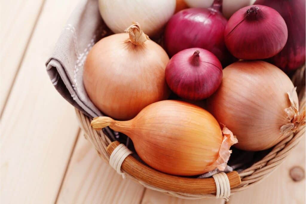 A woven basket holds an array of onions—white, yellow, and red—neatly arranged on a light wooden surface. Trick your way out of crying as you handle the cut onions with ease, transforming any kitchen task into a tear-free experience.