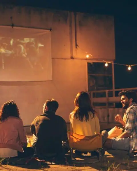 Five people sit on the ground outdoors at night, enjoying a fun movie night as they watch a film projected onto a screen, with string lights hanging nearby.