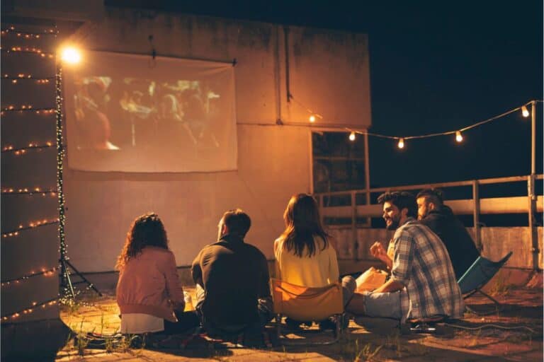 Five people sit on the ground outdoors at night, enjoying a fun movie night as they watch a film projected onto a screen, with string lights hanging nearby.