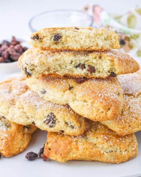 A stack of easy and delicious scones with raisins, dusted with powdered sugar, placed on a white plate. One scone is broken, showing a light, fluffy interior that makes this Bisquick recipe simply irresistible.