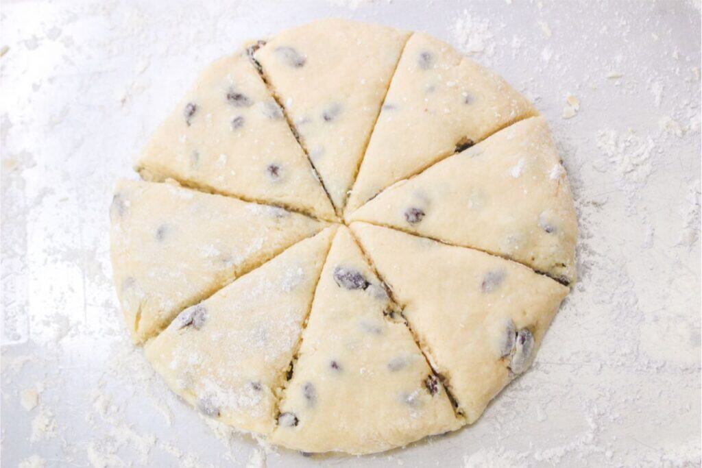 Unbaked scone dough with raisins, crafted from an easy and delicious Bisquick recipe, cut into eight triangular pieces and placed on a floured surface.