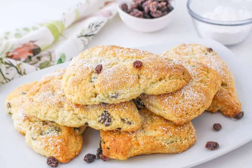 A plate of golden brown scones made with Bisquick is sprinkled with powdered sugar and topped with raisins. In the background, a bowl of raisins and a bowl of powdered sugar await your next recipe inspiration.