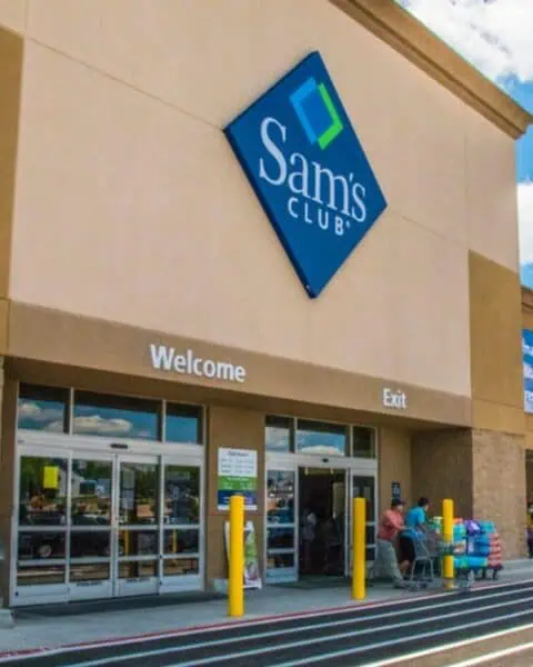 The exterior of a Sam's Club store with customers entering and exiting through the glass doors under a blue sign on a sunny day, where shoppers take advantage of exclusive Sam's Club Membership Deals.