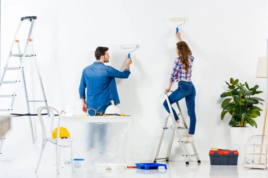 A man and woman are enhancing their space by painting a white wall with rollers. As the woman stands on a ladder, surrounded by painting supplies, a plastic-covered table, and a potted plant, they demonstrate simple ways of home improvement.