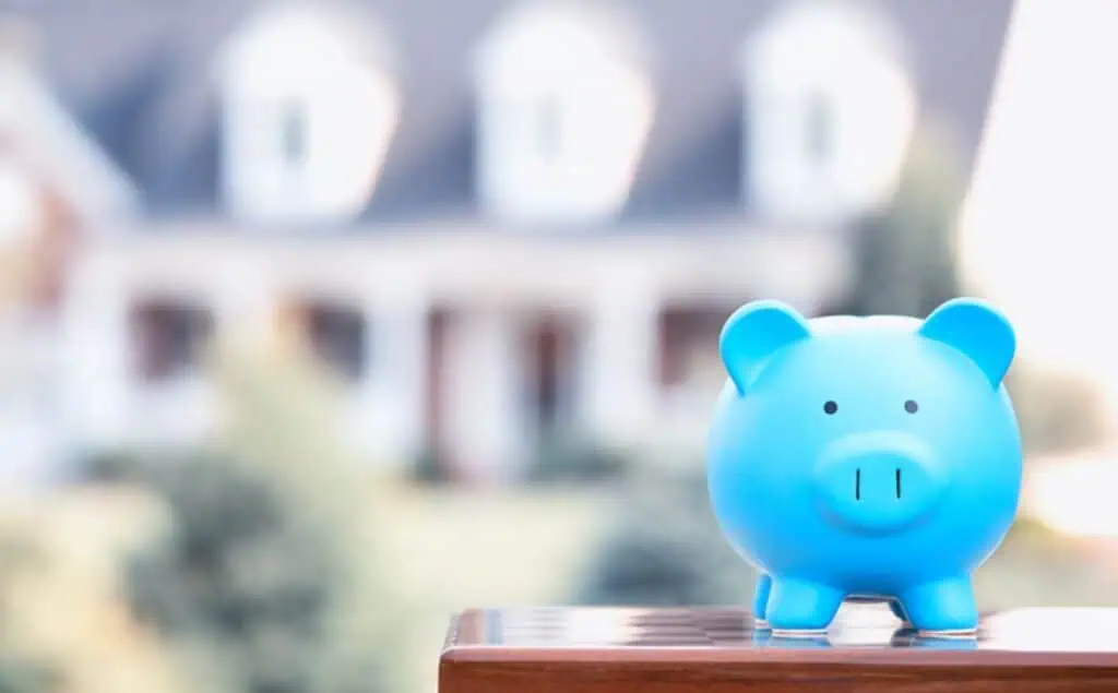 Blue piggy bank on a wooden surface, saving money for home improvement, with a blurred house in the background.