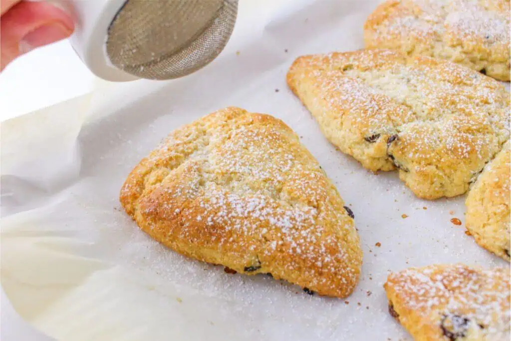 A hand gently sprinkles powdered sugar over freshly baked triangular scones on parchment paper, showcasing an easy scone recipe perfect for any occasion.