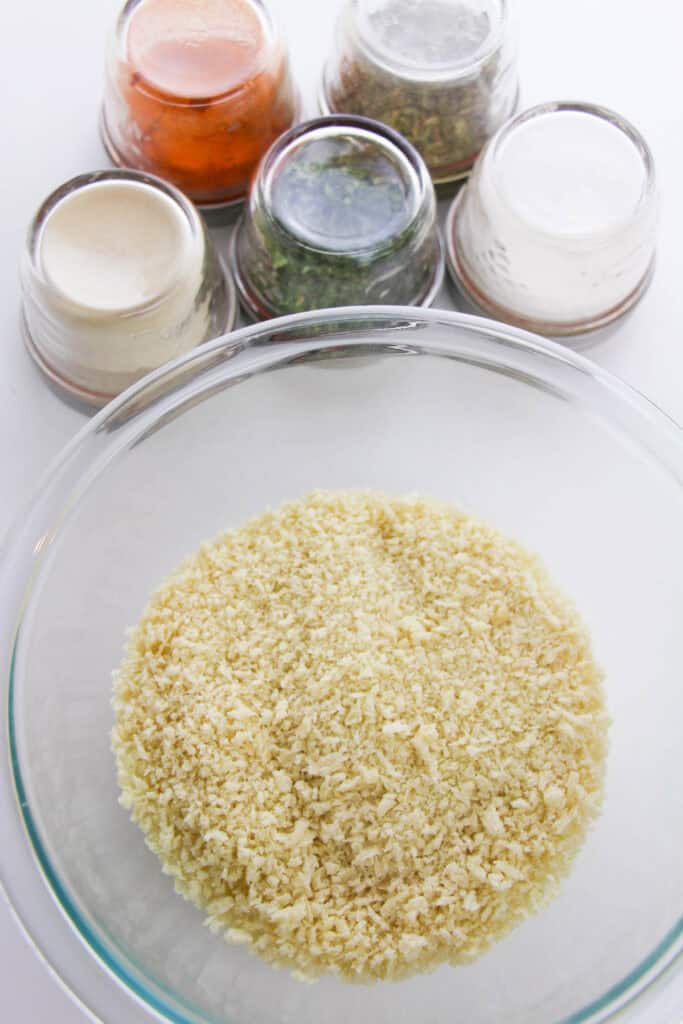 A clear bowl brimming with panko breadcrumbs sits in the foreground, ready for a homemade shake and bake adventure, while five small glass bowls with various spices linger invitingly in the background.