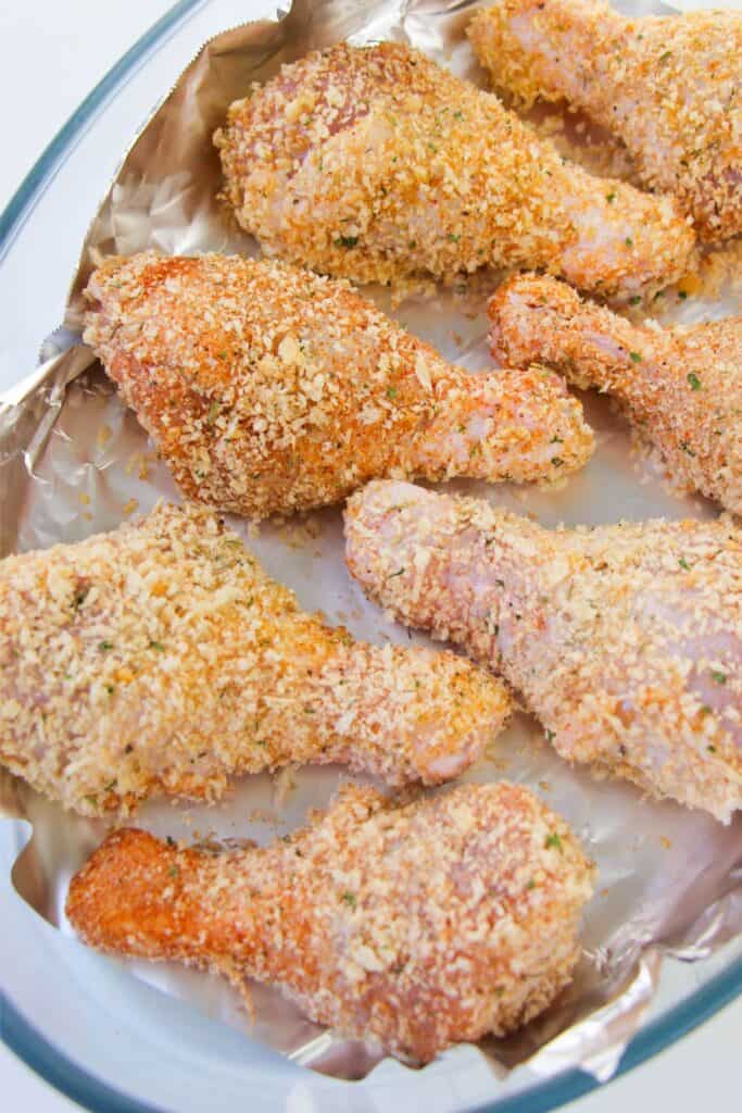 Raw breaded chicken drumsticks, seasoned with a homemade shake and bake mix, rest on a foil-lined baking tray, eagerly awaiting the oven.
