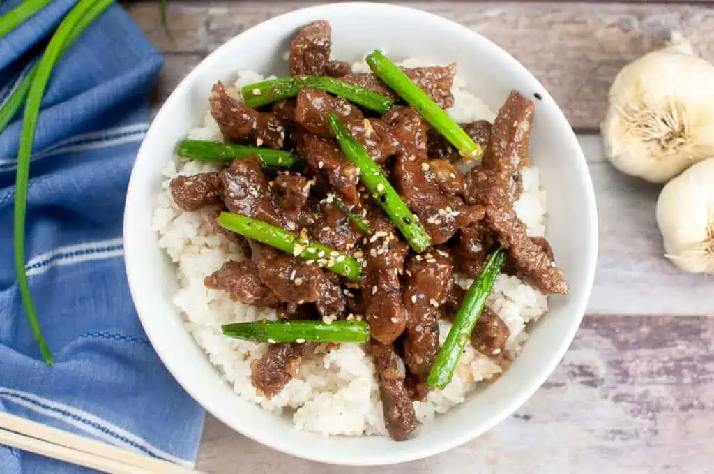 A bowl of Mongolian Beef stir-fry with green beans and sesame seeds over white rice sits on a wooden table, surrounded by garlic and a blue cloth. Perfect for those looking to discover a flavorful recipe.