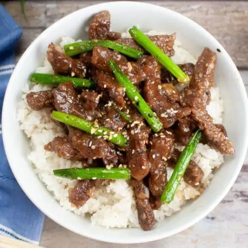 A bowl of Mongolian Beef stir-fry with green beans and sesame seeds over white rice sits on a wooden table, surrounded by garlic and a blue cloth. Perfect for those looking to discover a flavorful recipe.
