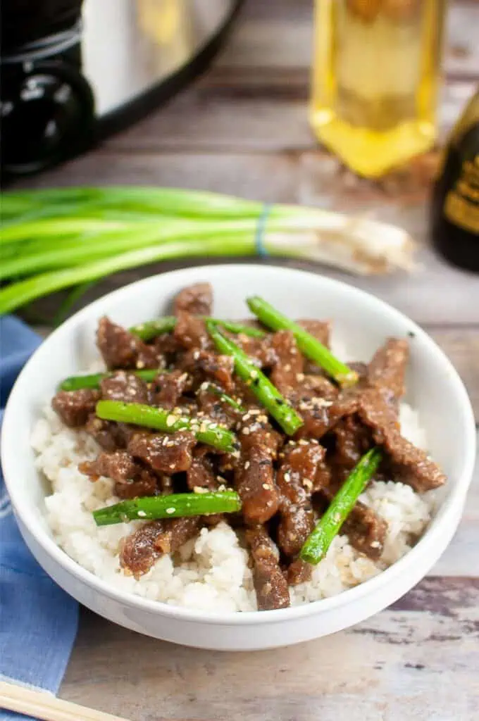 A bowl of rice topped with succulent Mongolian beef and green onions, garnished with sesame seeds. A bottle and extra green onions are in the background, offering a glimpse of the delightful recipe you can easily create at home.