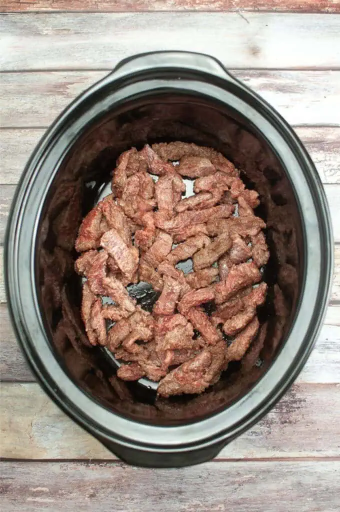 Slices of seasoned Mongolian beef nestled inside a black slow cooker, sitting on a wooden surface.