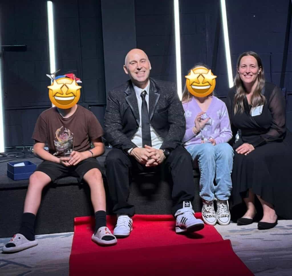 Four people sit on a platform against a dark background with vertical lights; two children's faces are covered with smiley face emojis.