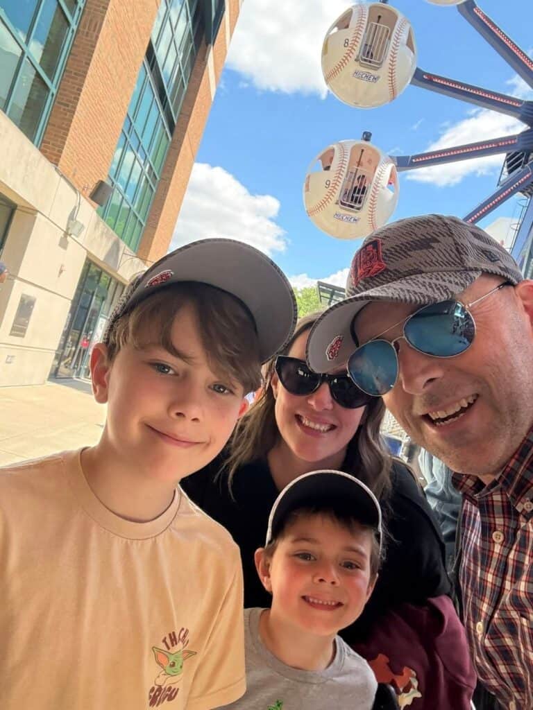 A group of four people wearing baseball caps and Stadium Dogs pose for a selfie outside, with oversized baseball decorations and a building in the background.