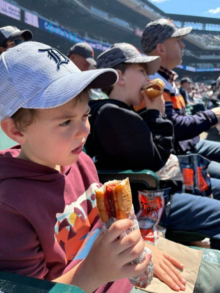Two boys and a man sit in stadium seats, wearing baseball caps and enjoying Stadium Dogs during a daytime baseball game.