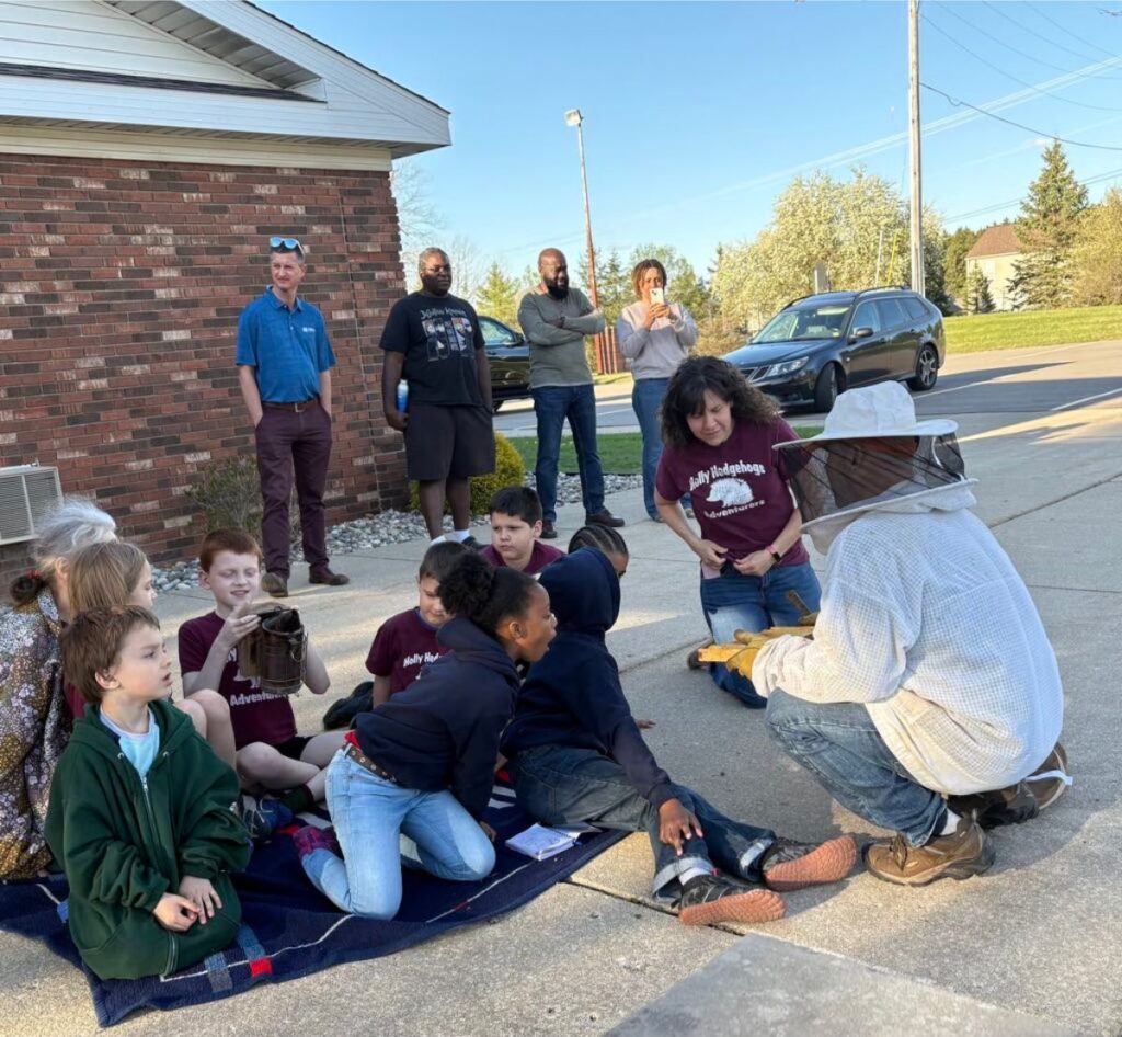A group of children and adults enjoy a sweet spring update as they watch a beekeeper in protective gear demonstrating equipment on a driveway; some adults stand in the background near parked cars and a brick building.