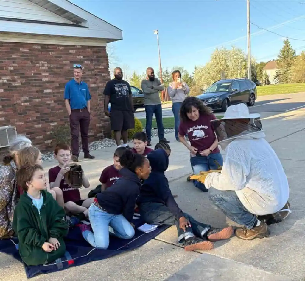 A group of children and adults enjoy a sweet spring update as they watch a beekeeper in protective gear demonstrating equipment on a driveway; some adults stand in the background near parked cars and a brick building.
