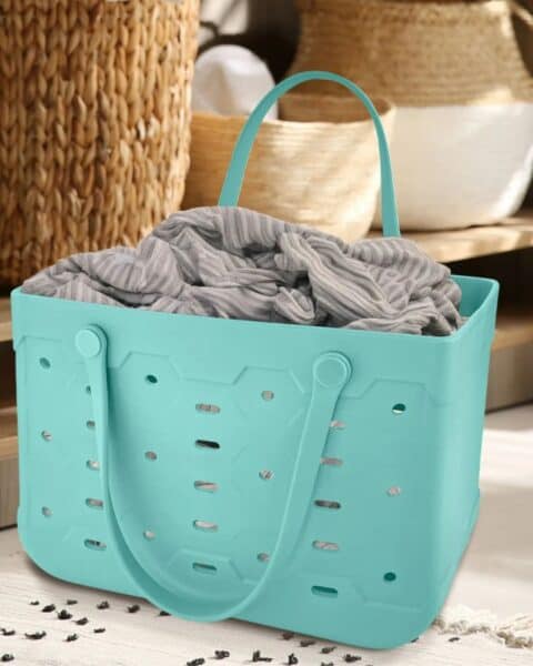 A teal plastic laundry basket with holes and handles, reminiscent of a Free Bogg Bag Dupe, holds a gray and white striped fabric, placed on a rug in a room with woven baskets in the background.