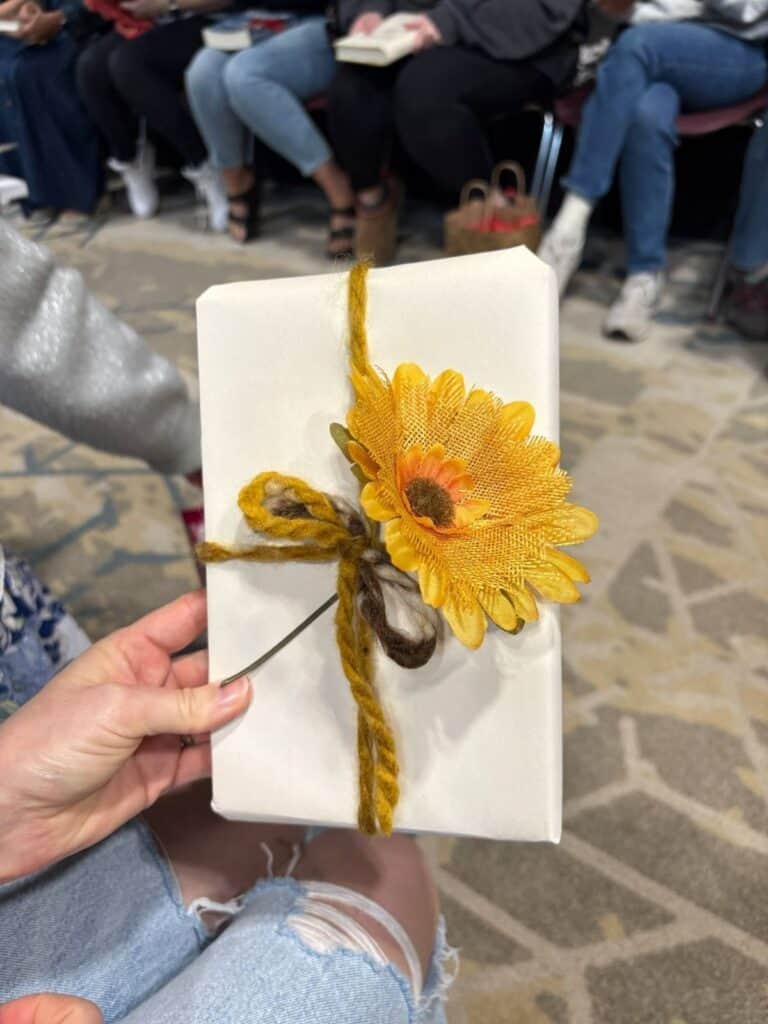 A hand holds a white-wrapped gift tied with yellow and brown yarn, topped with a large artificial yellow flower. People are seated in the background, capturing a warm moment for this Personal Post.