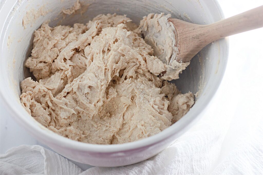 A mixing bowl filled with raw dutch oven bread dough being stirred with a wooden spoon.