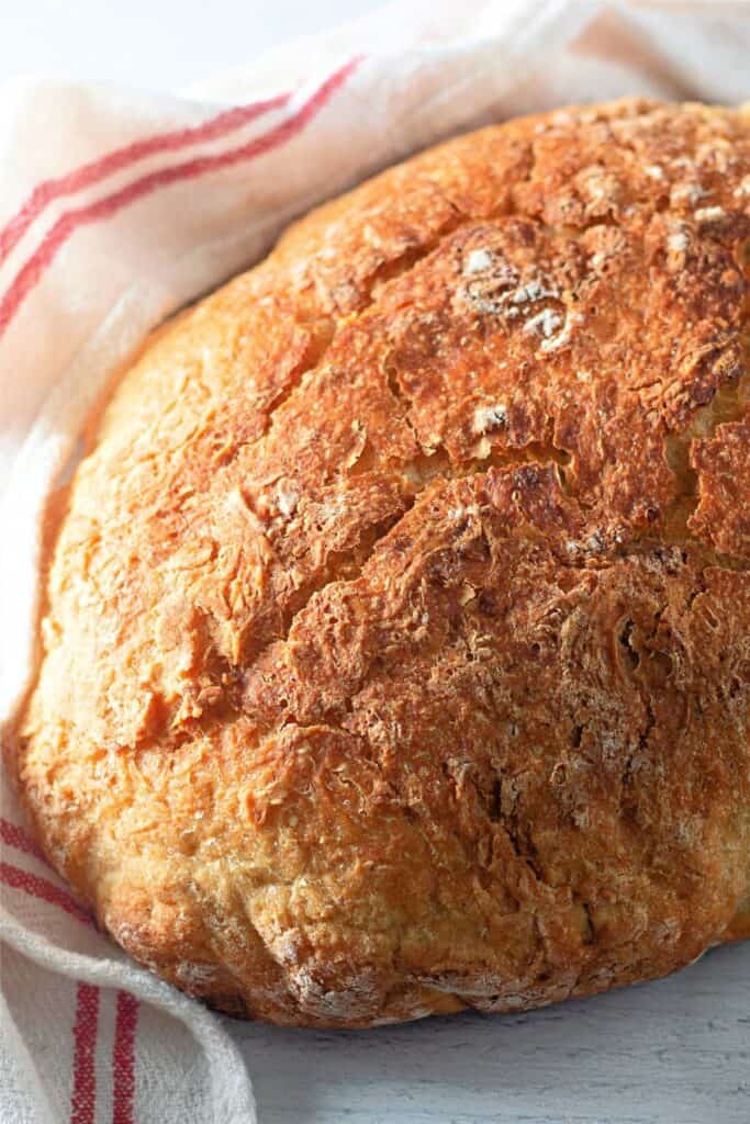 A round loaf of crusty Dutch oven bread rests on a white surface, partially wrapped in a white cloth with red stripes.
