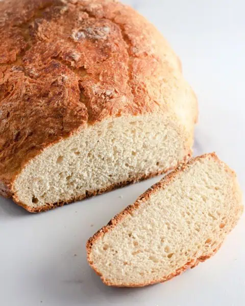 A loaf of rustic Dutch oven bread with a golden crust is shown on a white surface, with one slice cut and placed in front of the loaf.
