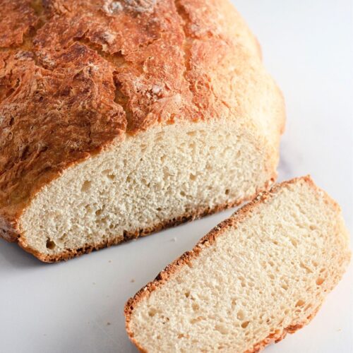 A loaf of rustic Dutch oven bread with a golden crust is shown on a white surface, with one slice cut and placed in front of the loaf.