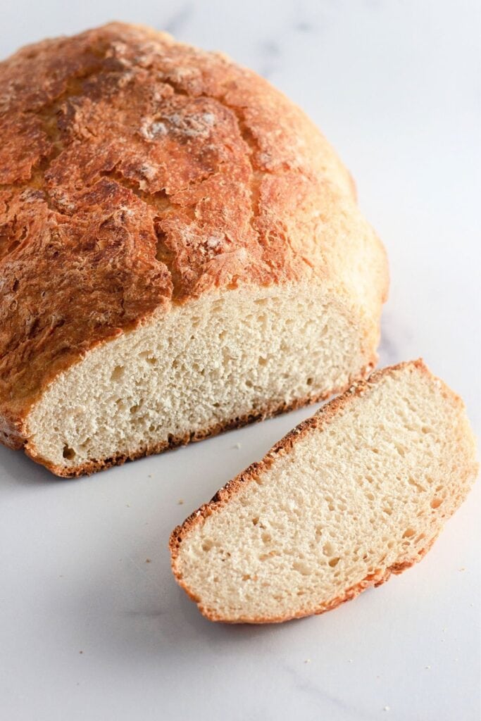 A loaf of rustic Dutch oven bread with a golden crust is shown on a white surface, with one slice cut and placed in front of the loaf.