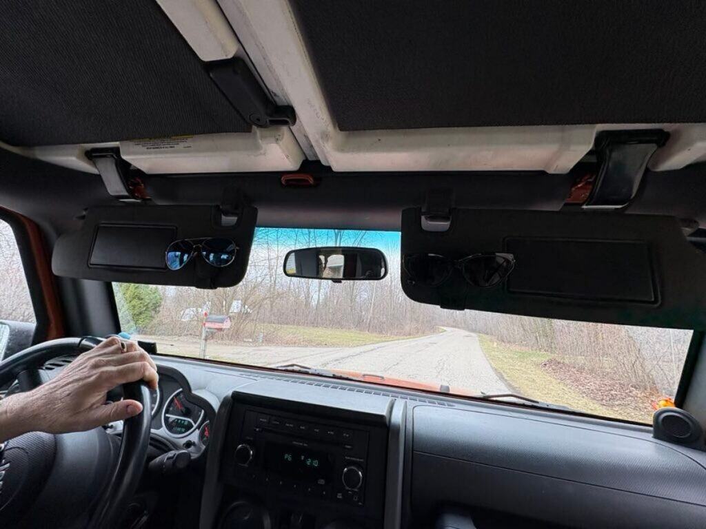 Interior view of a vehicle from the driver's seat, showing two pairs of sunglasses on sun visors, a hand on the steering wheel, and a road ahead lined with trees—perfect for a family trip or visiting four generations together.