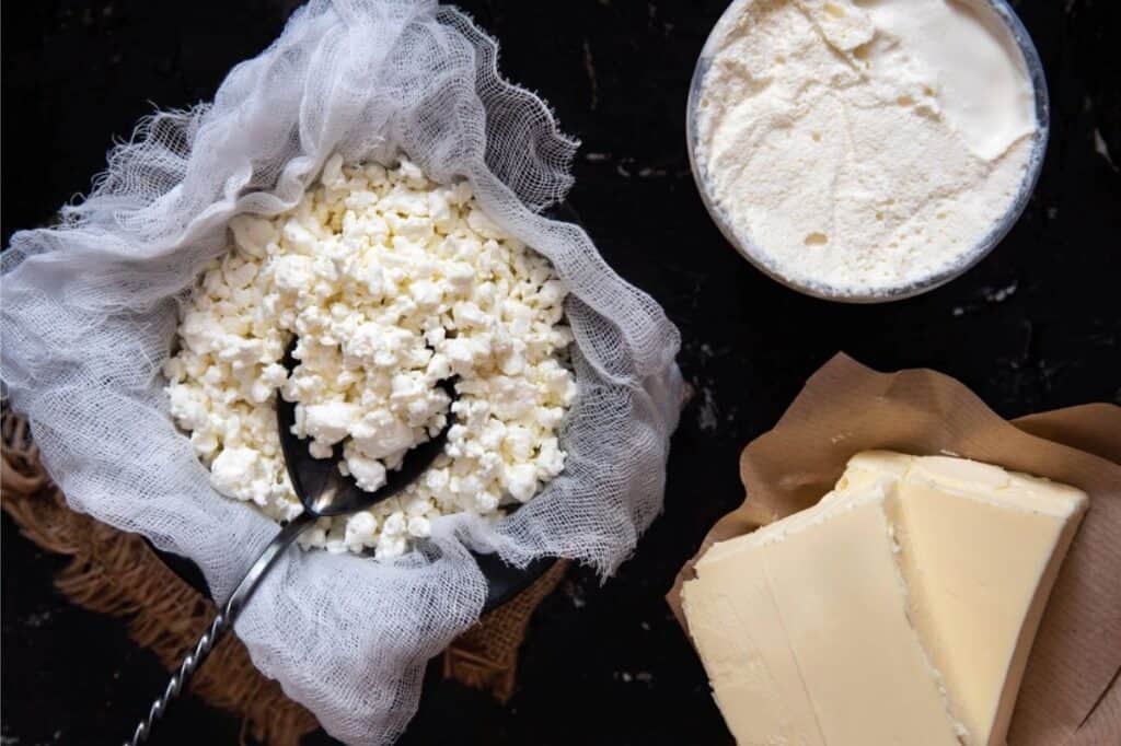 A bowl of homemade cheese with a spoon, a block of butter on parchment paper, and a bowl of cream are arranged on a dark surface.