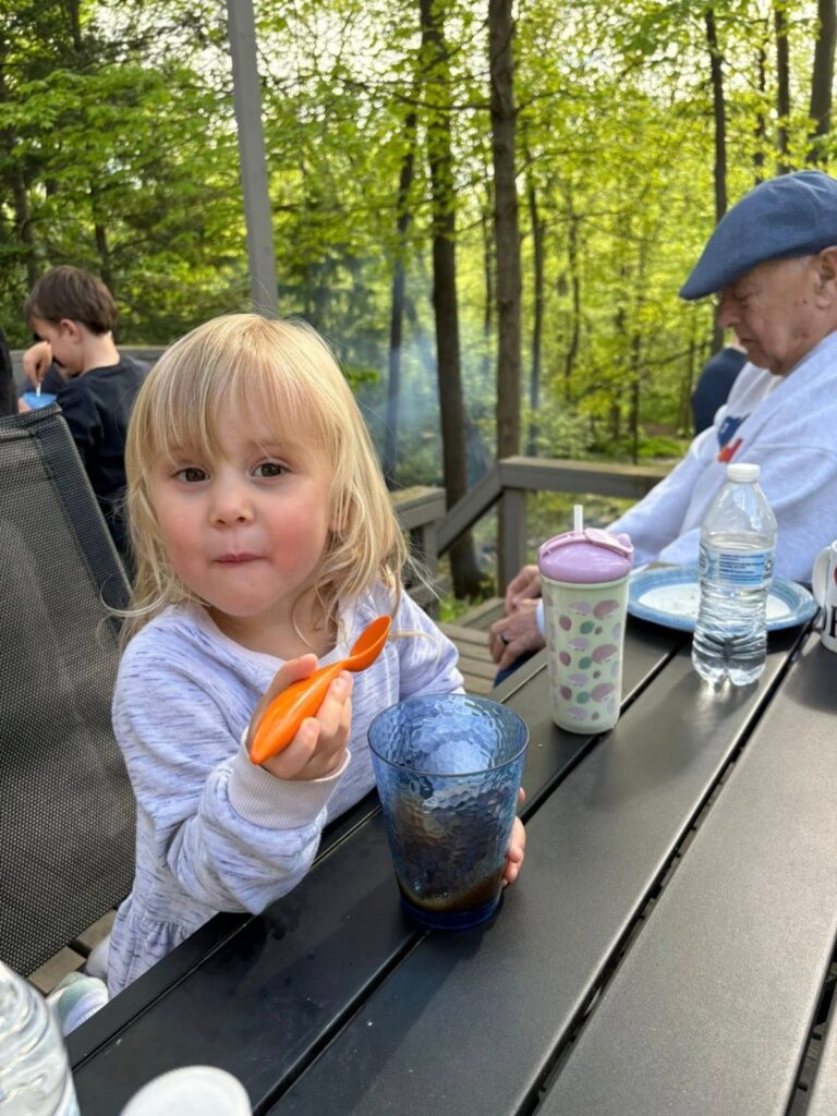 A young child with sticker earrings sits at an outdoor table holding a spoon and cup, with drinks and water bottles nearby; adults are seated in the background, surrounded by trees.