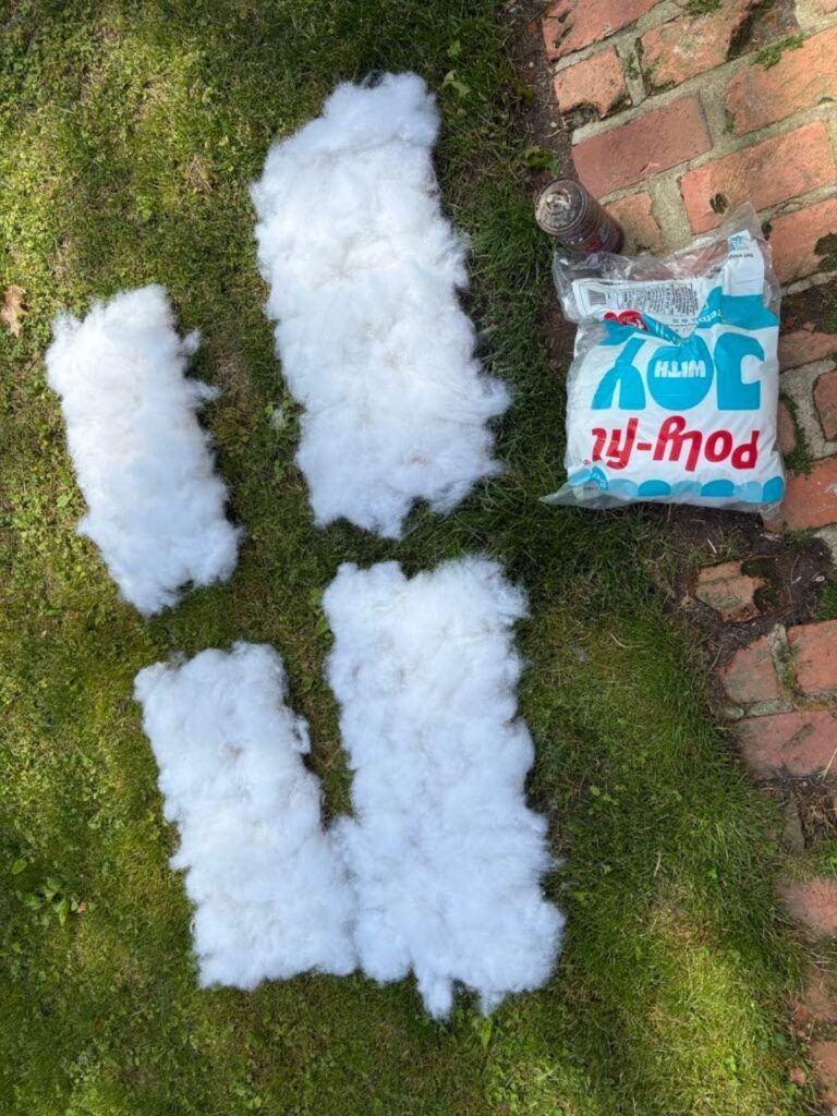 Four sections of white polyester fiberfill are spread on grass next to an open bag of Poly-fil stuffing, a plastic bottle, and a set of Sticker Earrings resting on a brick path.
