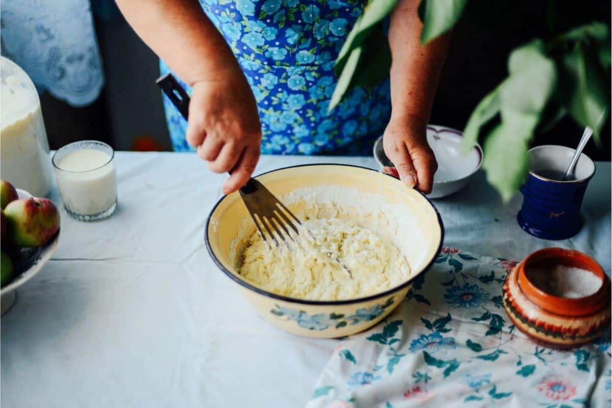 A person in a blue floral dress mixes dough in a large bowl on a table set with various kitchen items—examples of life skills like cooking and preparing ingredients, surrounded by a glass of milk, apples, and a patterned cloth.