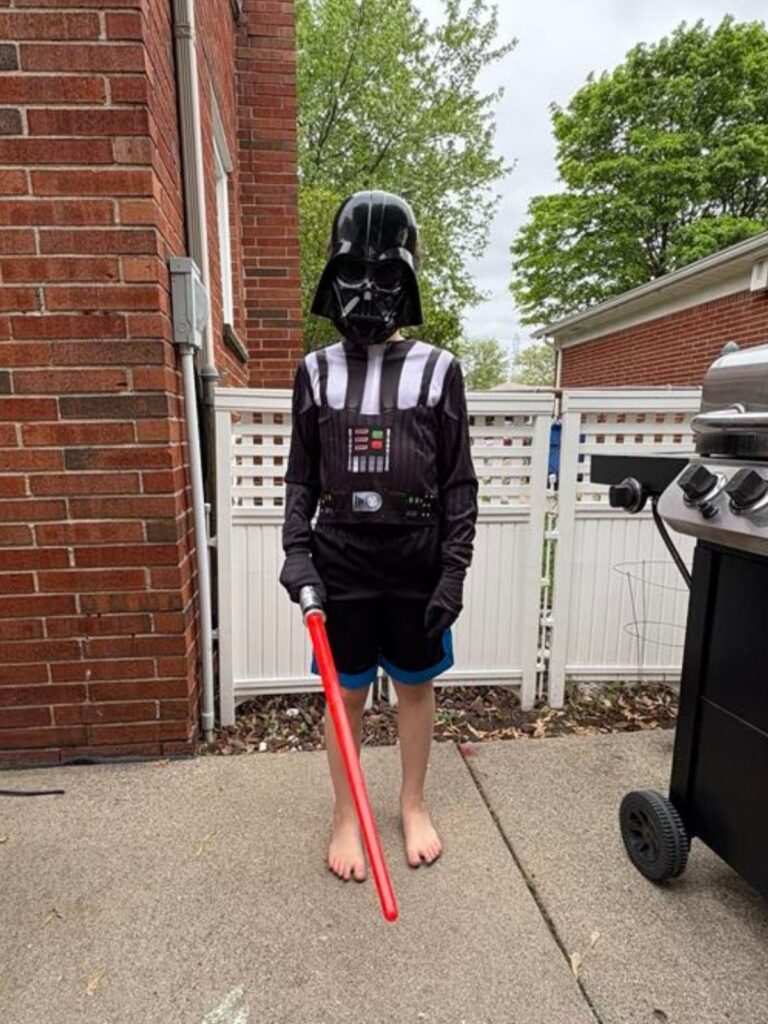 A child wearing a Darth Vader costume with a red lightsaber stands on a patio beside a grill, as seen in this NEW Update, with a white fence and brick buildings in the background.