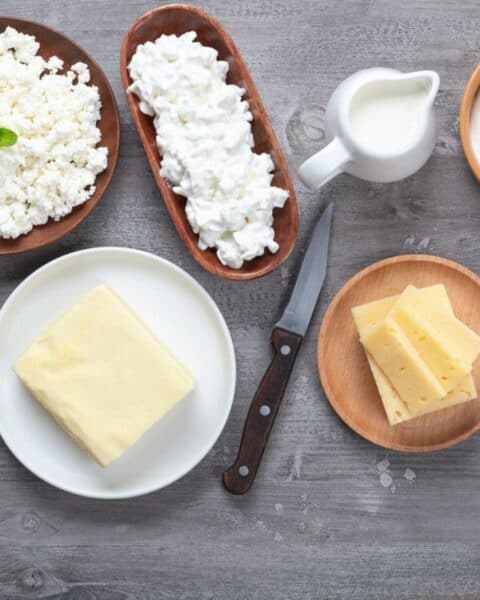 Assorted dairy products—including cottage cheese, cream, milk, sliced cheese, and a block of butter—are displayed on a wooden surface with a knife and spoon, perfect for exploring how to make cheese at home.
