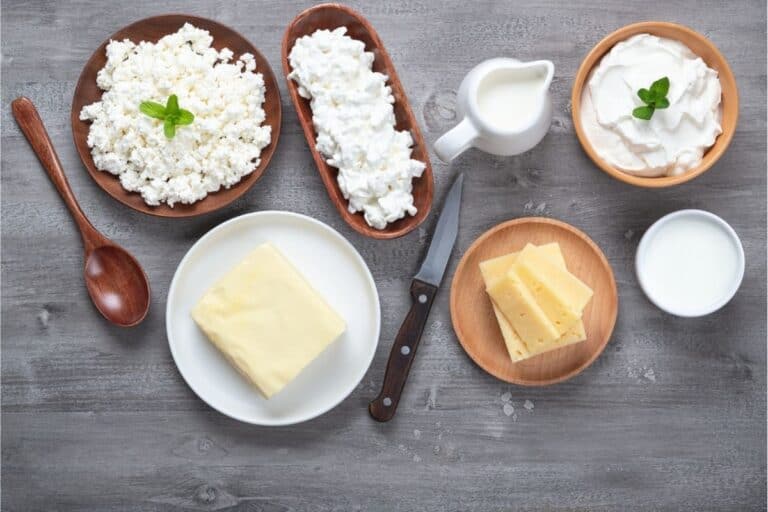Assorted dairy products—including cottage cheese, cream, milk, sliced cheese, and a block of butter—are displayed on a wooden surface with a knife and spoon, perfect for exploring how to make cheese at home.