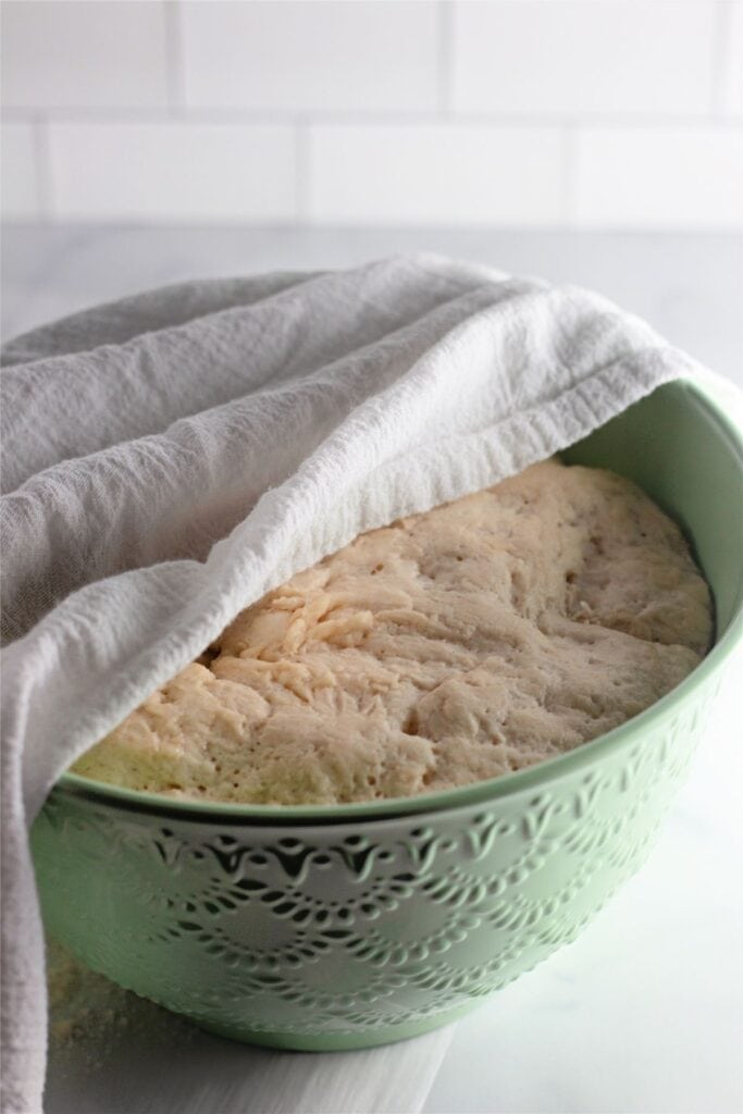 A green textured bowl filled with rising dutch oven bread dough is partially covered by a white cloth on a white countertop.