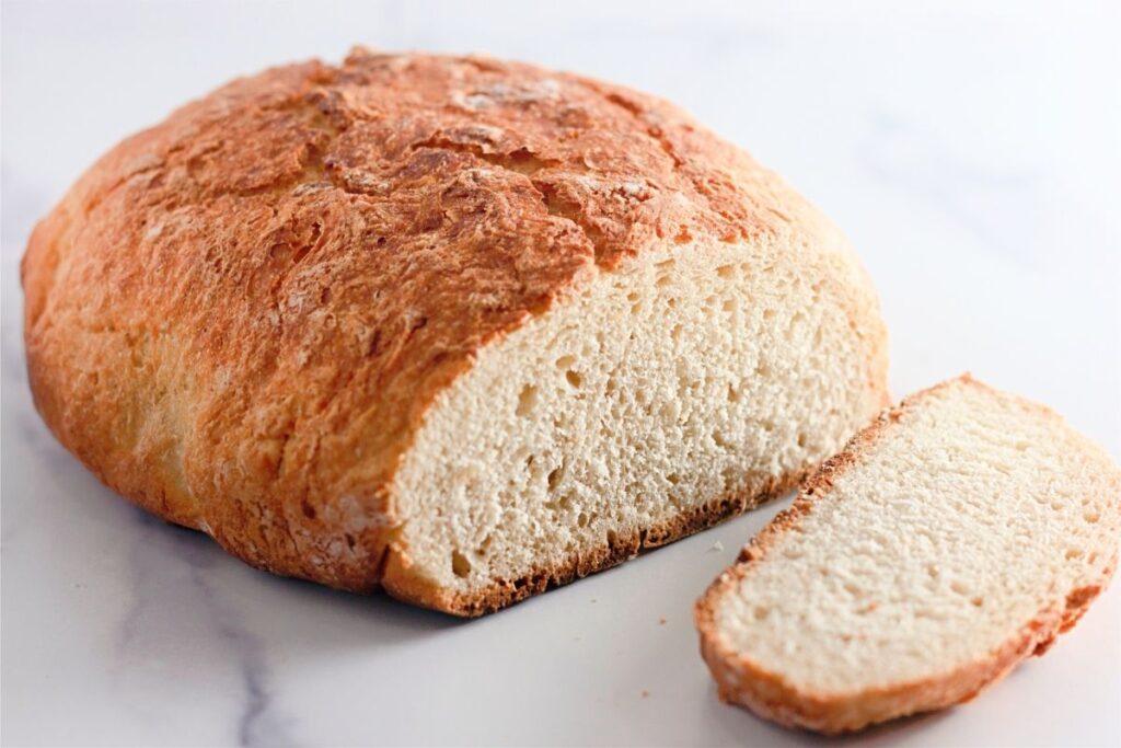 A round loaf of crusty dutch oven bread with one slice cut and placed in front on a white surface.