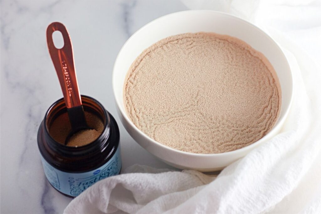A bowl filled with dry yeast sits next to an open jar of yeast with a measuring spoon inside, perfect for preparing dutch oven bread, all arranged on a white surface with a white cloth.