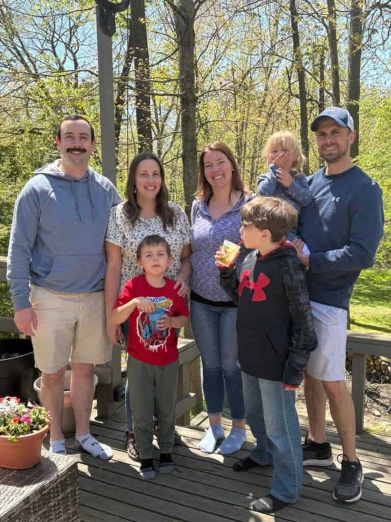 Three adults and four children stand together on a wooden deck outdoors, surrounded by trees, on a sunny day—capturing generations together, as some of the children hold snacks.