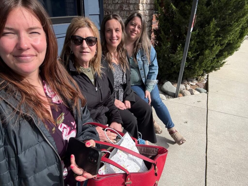 Four women sit closely together on an outdoor bench in sweet spring sunshine, smiling at the camera. One holds a red purse and smartphone. Trees and a sidewalk are visible, capturing a bright moment perfect for a spring update.