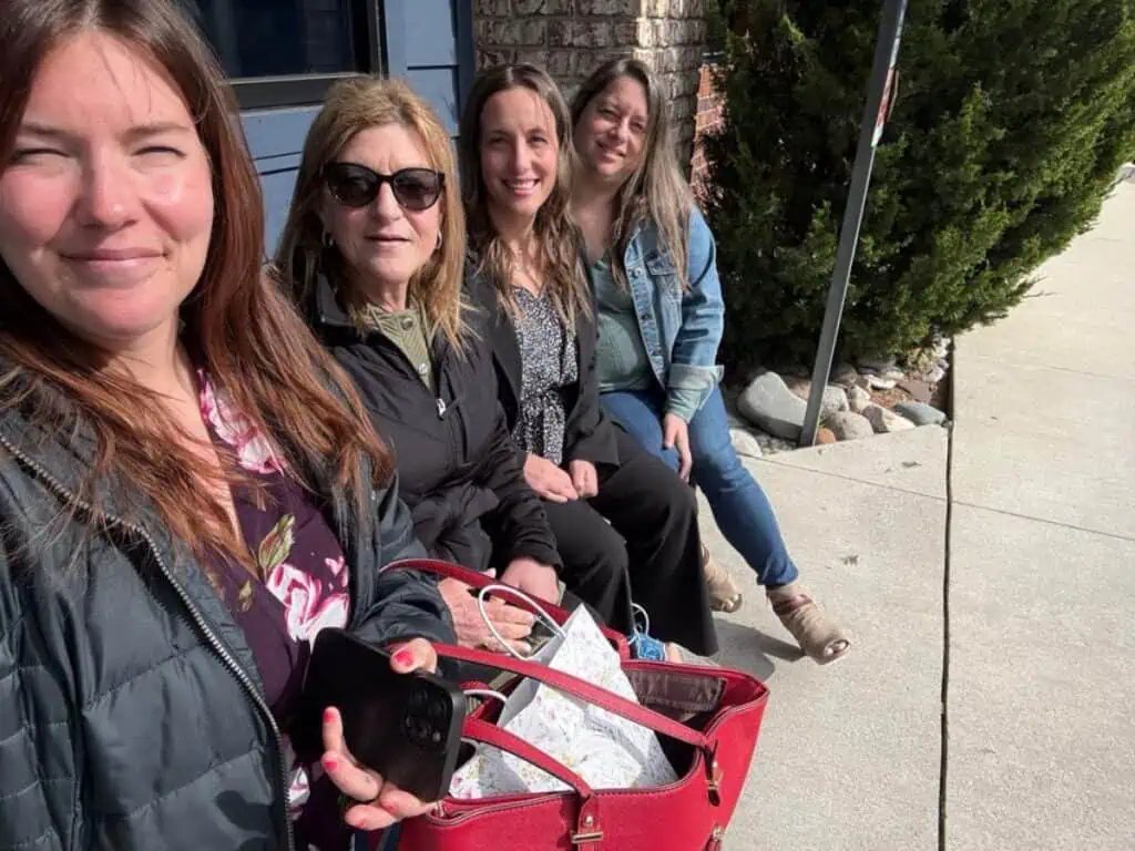 Four women sit closely together on an outdoor bench in sweet spring sunshine, smiling at the camera. One holds a red purse and smartphone. Trees and a sidewalk are visible, capturing a bright moment perfect for a spring update.