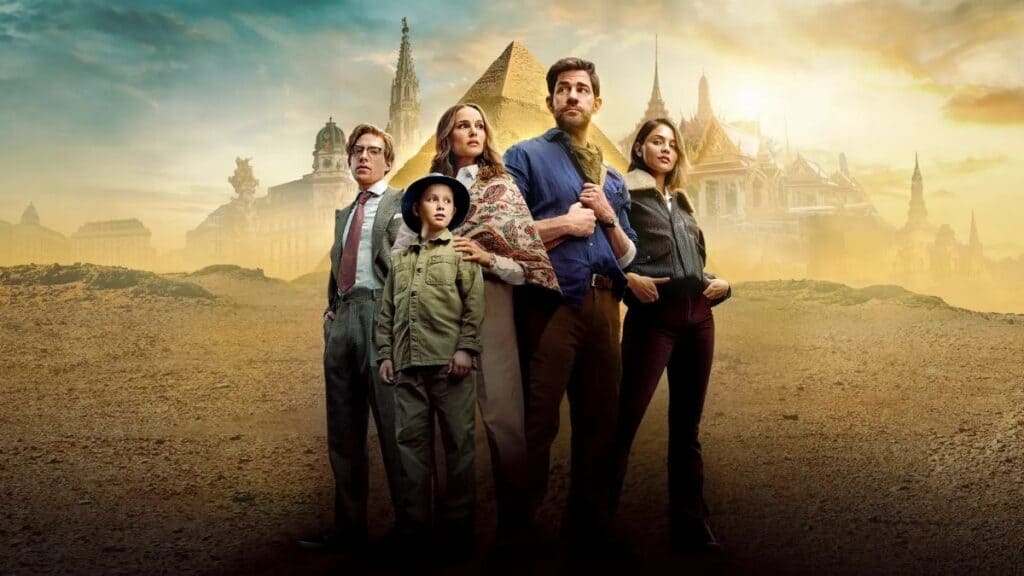 Five people stand together on sandy ground with architectural landmarks, including pyramids and temples, in the background under a cloudy sky. One wears playful sticker earrings, adding a modern touch to the ancient setting.