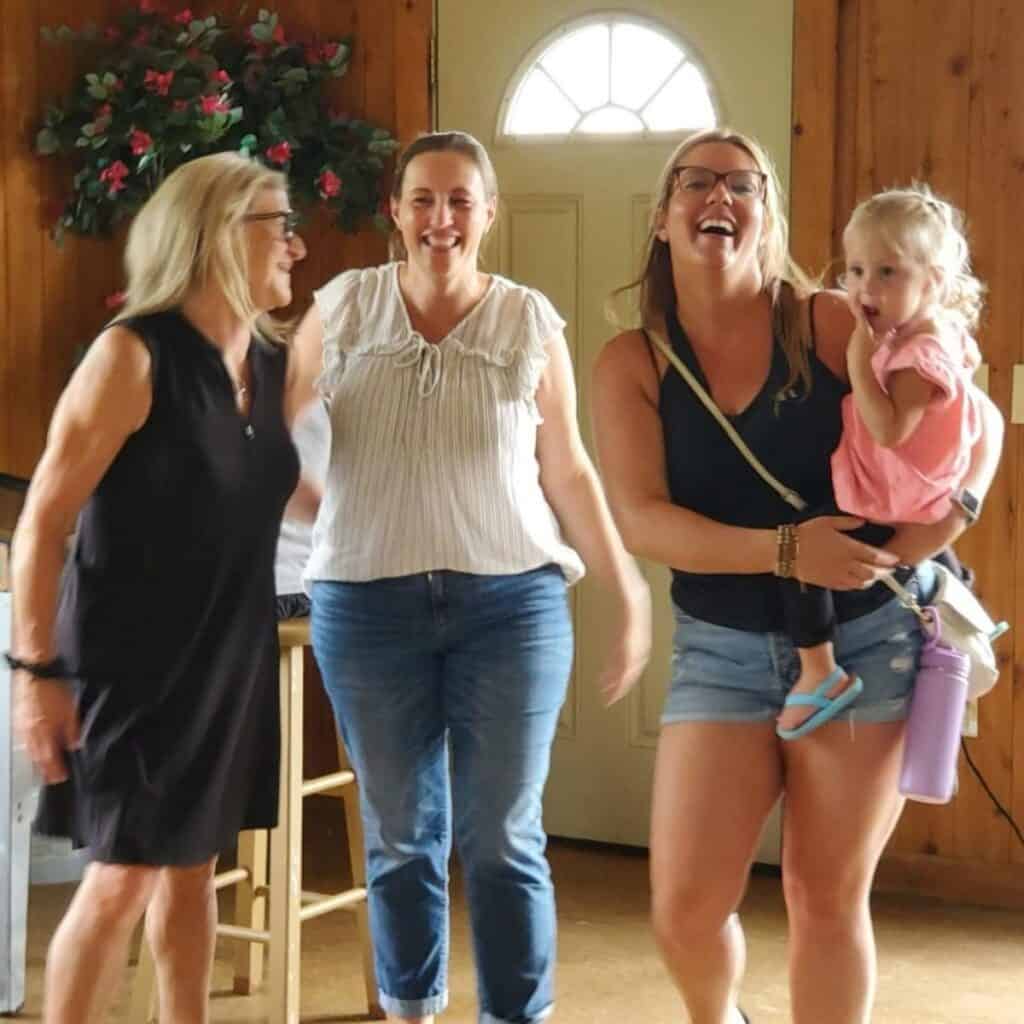 Three women stand indoors smiling, one holding a young child on her hip—four generations together. A wooden wall, door, and potted plant reflect the warmth often found on a family farm.