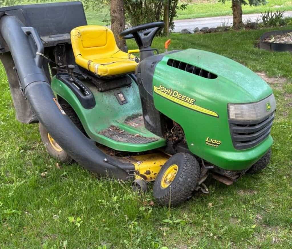 A green John Deere L100 riding lawn mower with a yellow seat and attached grass collection tube is parked on grass near trees, reflecting the legacy of four generations devoted to family farming.