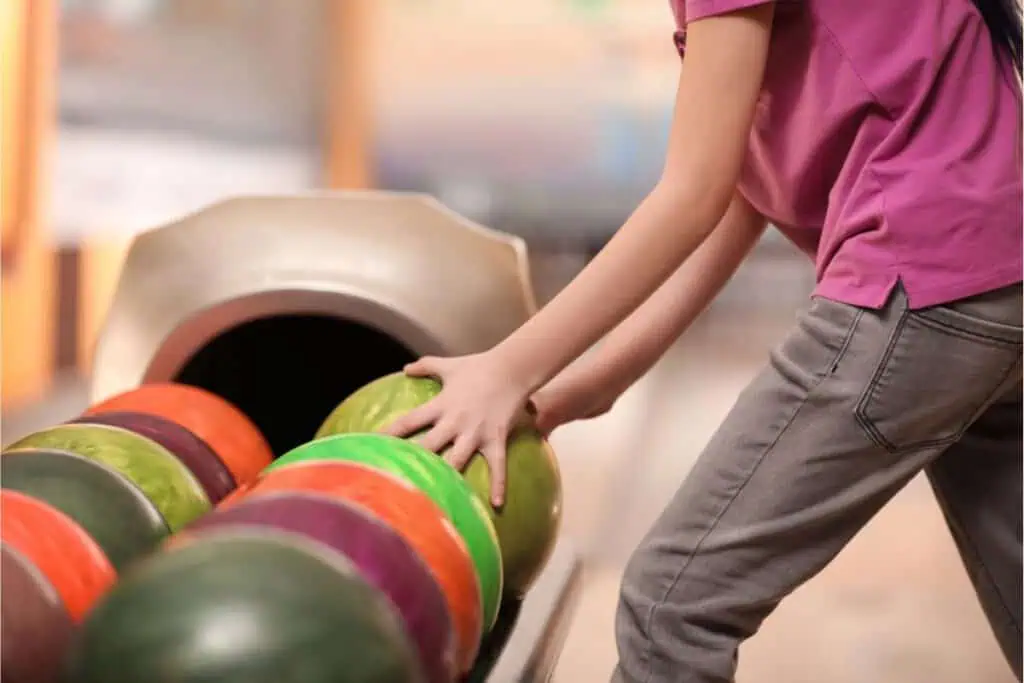 A person wearing a pink shirt and gray pants selects a green bowling ball from a rack of colorful bowling balls at a kids bowl free event at the bowling alley.